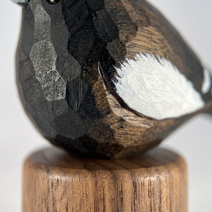 Close-up of a carved wooden Lark bunting figurine with dark black and brown wood grain, a white painted wing, and a round wooden base on a light gray background.
