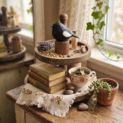 A rustic two-tier wooden serving tray styled on a table by a window, holding a black-and-white Lark bunting figurine, small bottles, dried flowers, keys, and stacked vintage books, alongside a cup, stones, and a potted trailing succulent.