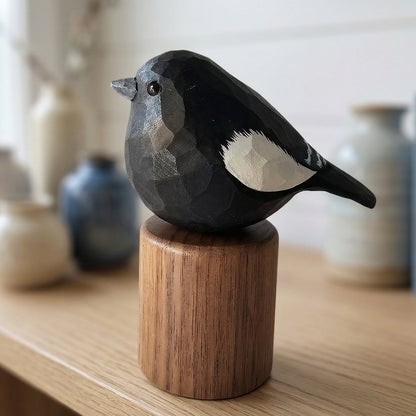 A hand-carved wooden Lark bunting figurine with a faceted geometric design, painted in black, gray, and white, mounted on a cylindrical wooden base and displayed on a light wood shelf.