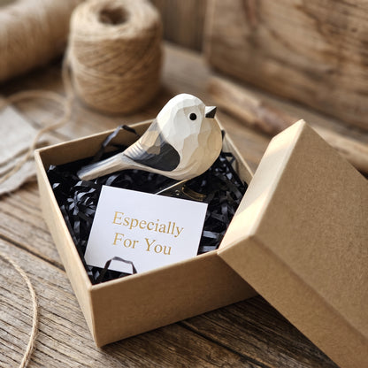 A hand-carved wooden Long-Tailed Tit clip-on bird ornament resting inside an open kraft paper gift box filled with black shredded paper, featuring a white body with carved black wing and tail accents, alongside a white card reading "Especially For You" in the foreground on a rustic wooden surface.