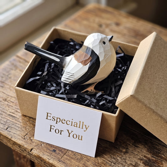 A hand-carved wooden Long-Tailed Tit clip-on bird ornament resting inside an open kraft paper gift box filled with black shredded paper, featuring a white head, a black back, a long dark tail, and a white card reading "Especially For You" placed in the foreground on a rustic wooden surface.