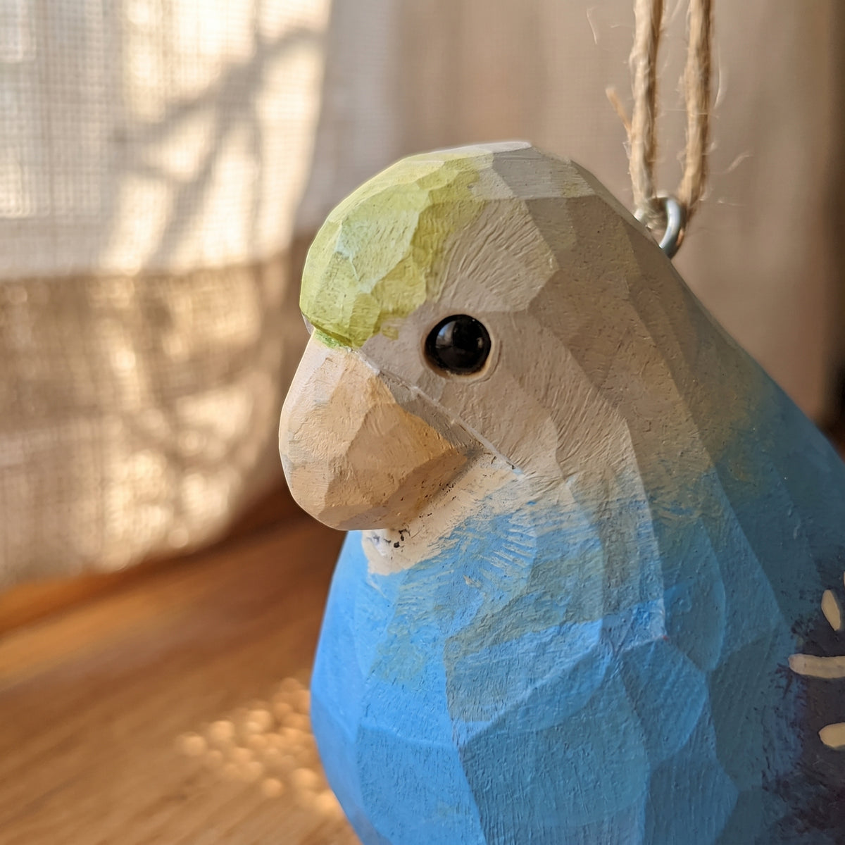 A close-up of a hand-carved wooden Lovebirds ornament, likely a parrot, featuring a faceted geometric design, painted in shades of blue, beige, and light green, with a natural jute hanging twine.