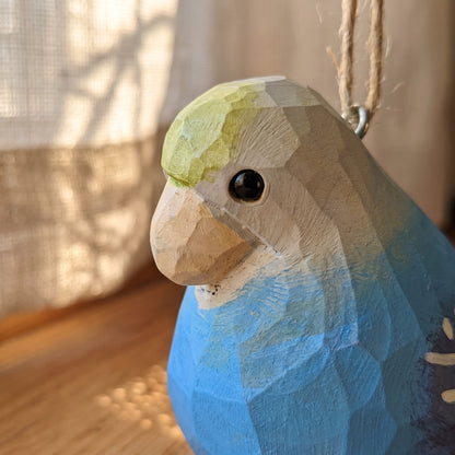 A close-up of a hand-carved wooden Lovebirds ornament, likely a parrot, featuring a faceted geometric design, painted in shades of blue, beige, and light green, with a natural jute hanging twine.