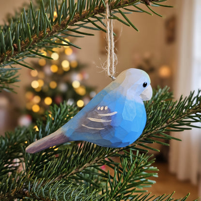 A handcrafted wooden Lovebirds ornament in shades of blue, gray, and white, hanging from a natural twine loop on a green pine tree branch with a blurred Christmas tree in the background.