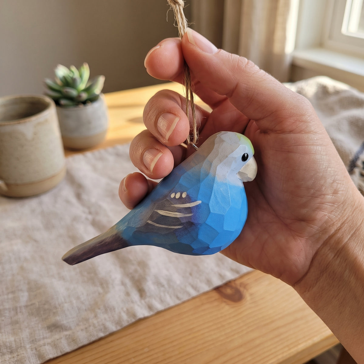 A hand holding a small wooden Lovebirds  ornament painted in shades of blue, gray, and white, with a natural wood hanging loop. The background shows a cozy indoor setting with a wooden table, linen cloth, and small potted plants.