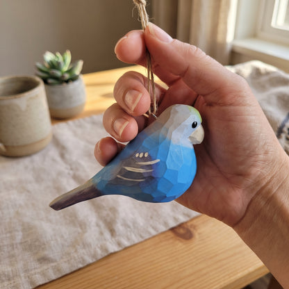 A hand holding a small wooden Lovebirds  ornament painted in shades of blue, gray, and white, with a natural wood hanging loop. The background shows a cozy indoor setting with a wooden table, linen cloth, and small potted plants.