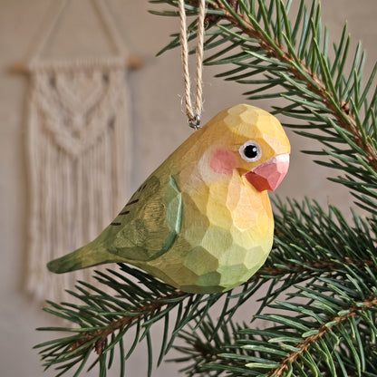 A hand-carved wooden Lovebirds ornament with a geometric faceted finish, hanging from a jute string on a pine tree branch. The bird is yellow and green with a pink beak and pink cheek accent, set against a softly blurred background with a macrame wall hanging.