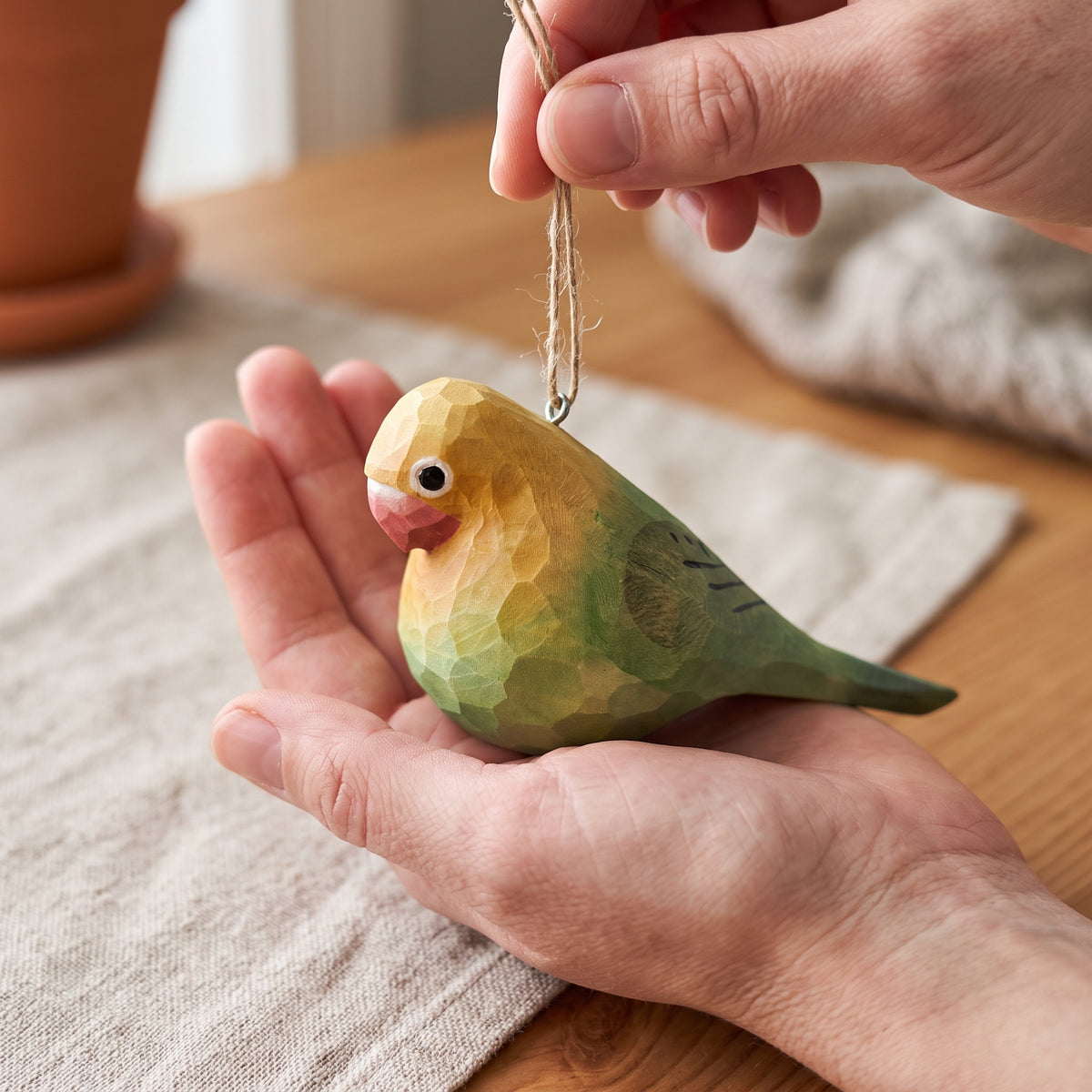 A hand holds a small carved Lovebirds ornament in the palm of another hand. The ornament has a faceted, geometric texture and is painted yellow, green, and blue, with a pink beak and a small black-and-white eye.