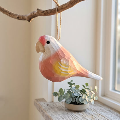 A faceted wooden Lovebirds ornament, resembling a cockatoo, hanging from a natural branch. The ornament features a geometric, low-poly design with painted colors of pink, yellow, and white.