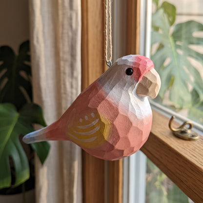 A low-poly wooden Lovebirds ornament, resembling a pink and white cockatoo with a yellow accent on its wing, hanging from a rope hook in front of a window with plants in the background.