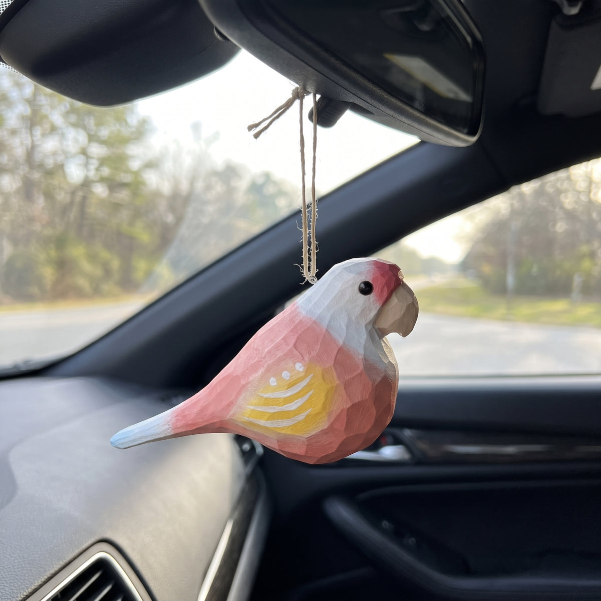 A pink and white Lovebirds-shaped car air freshener with a yellow patterned wing, hanging from a car's rearview mirror.
