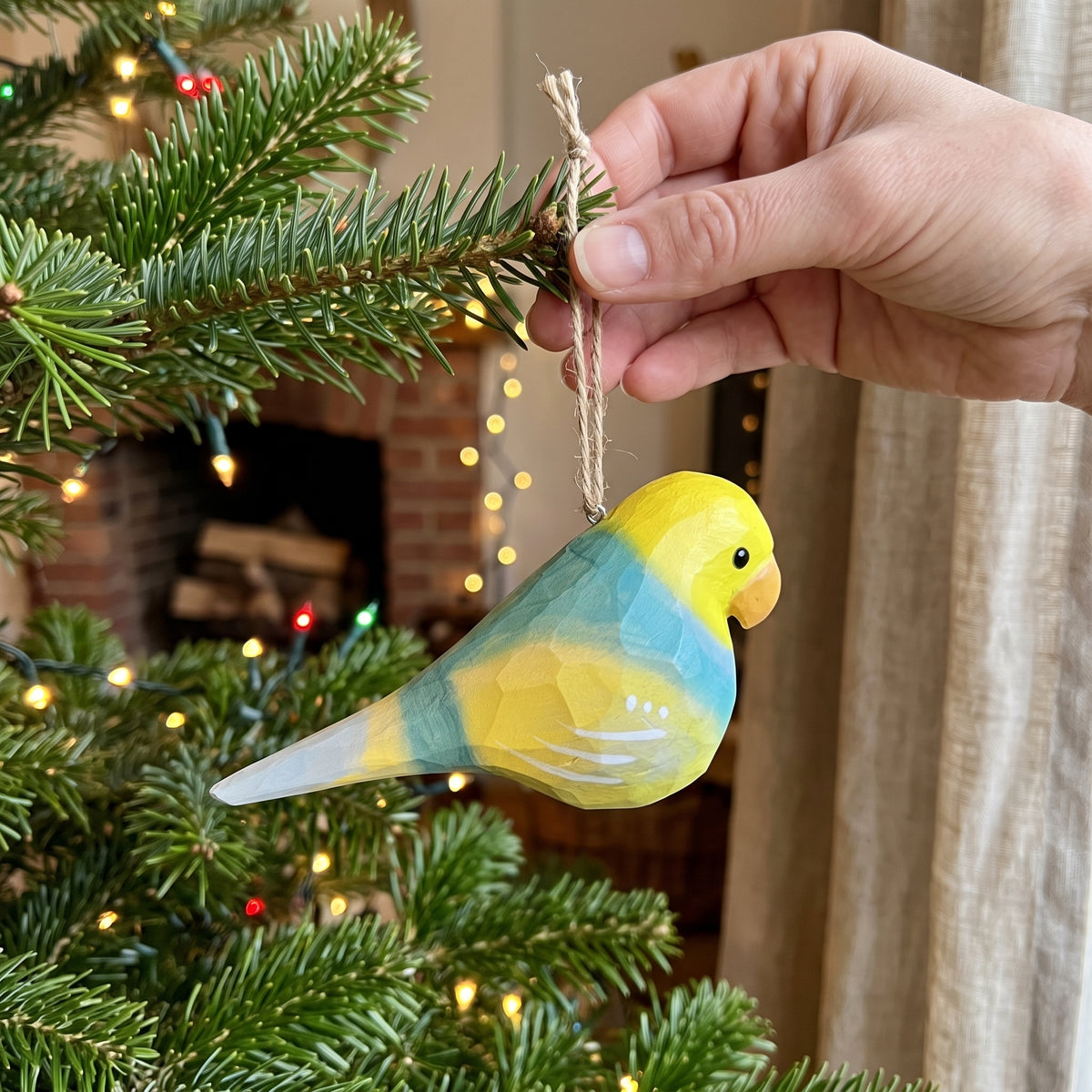 A hand hangs a small Lovebirds-shaped Christmas ornament from a pine tree branch. The ornament is yellow and light blue with a small orange beak and black eye, set against a softly blurred indoor background with warm lights.