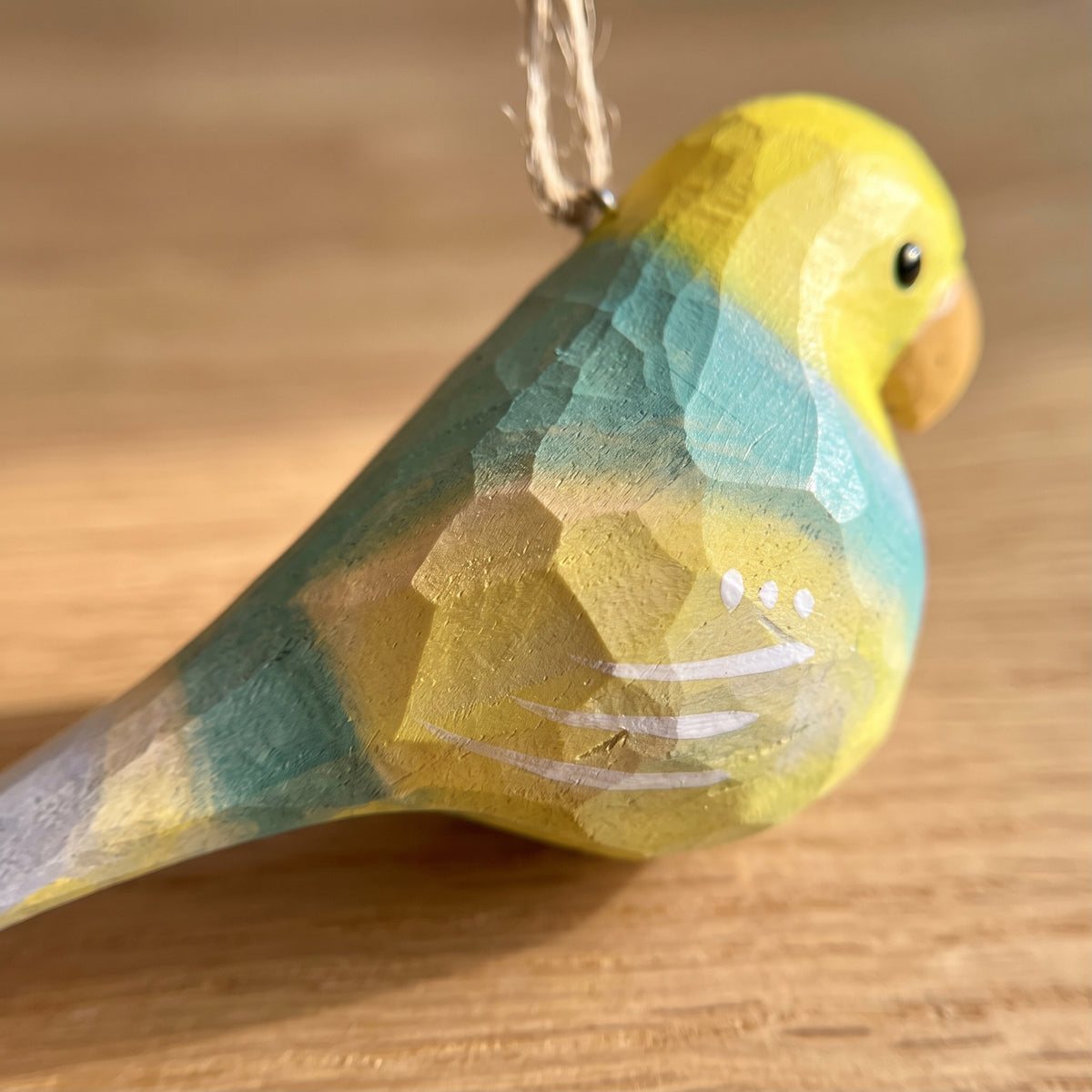 A close-up of a handcrafted wooden Lovebirds ornament painted in yellow and teal, with a small hanging twine loop at the top, set against a warm wooden background.