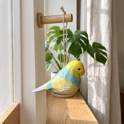 A small hanging Lovebirds ornament in yellow, blue, and white hangs from a wooden dowel mounted on a window frame, with a potted plant and curtain in the background.