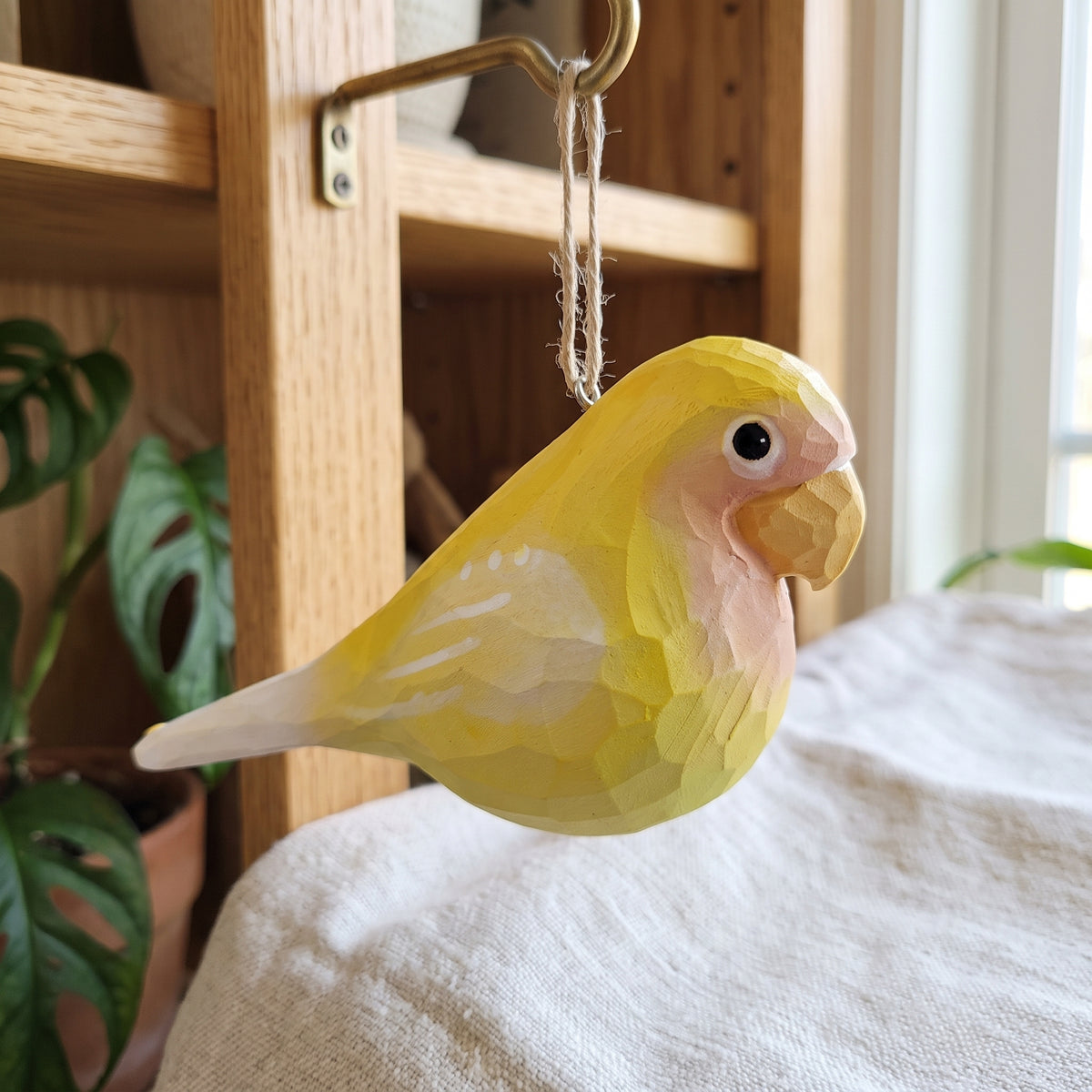 A handcrafted wooden Lovebirds ornament, shaped like a yellow lovebird, hanging from a brass hook on a wooden shelf in a home setting.