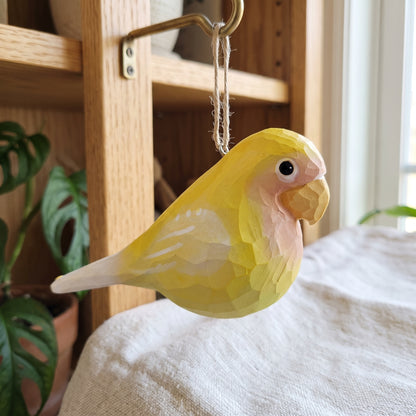 A handcrafted wooden Lovebirds ornament, shaped like a yellow lovebird, hanging from a brass hook on a wooden shelf in a home setting.