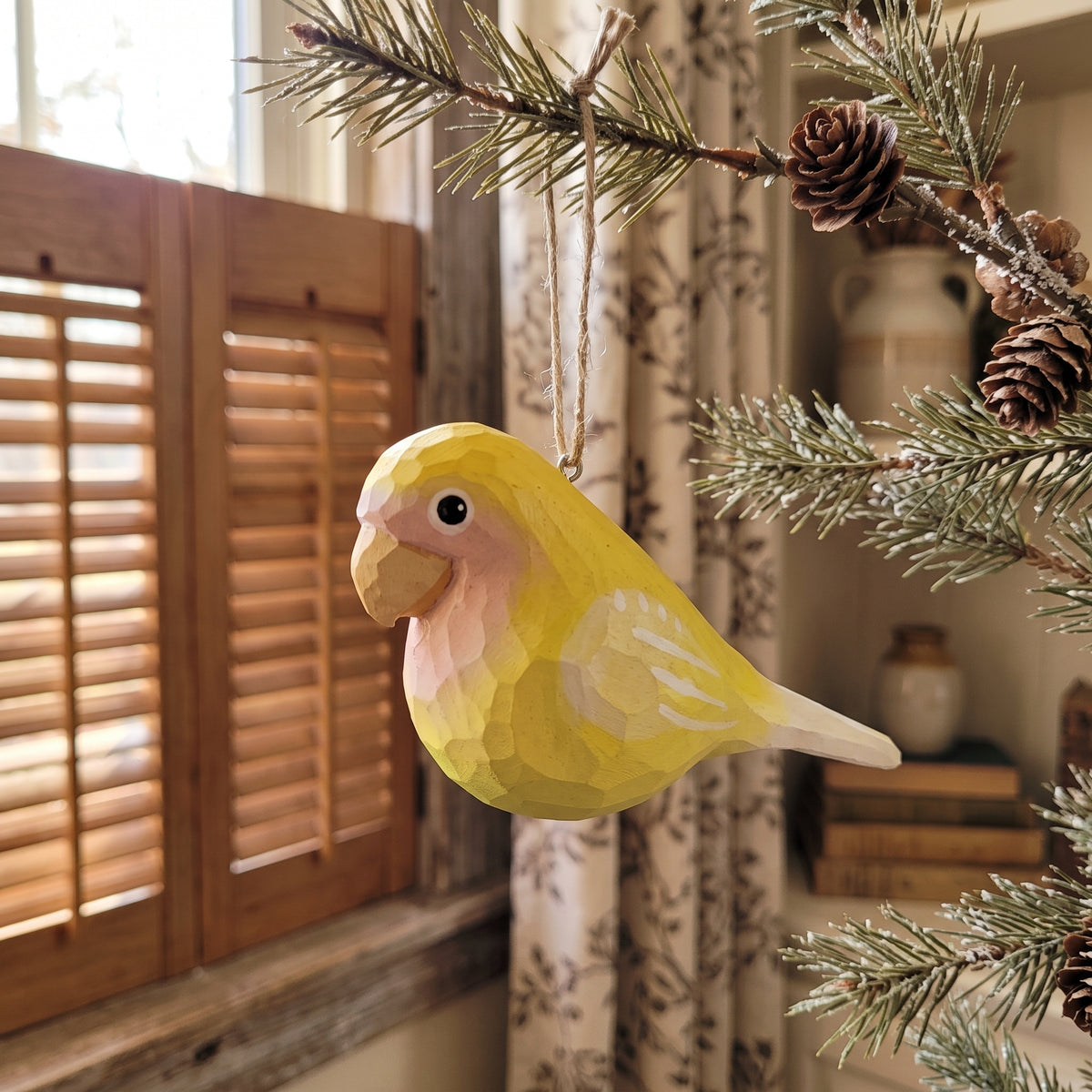 A yellow Lovebirds-shaped Christmas ornament hanging from a pine branch in a cozy indoor setting with wooden shutters and curtains in the background.