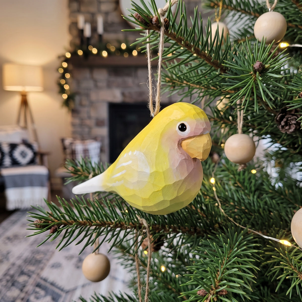 A yellow Lovebirds-shaped Christmas ornament hanging on a pine tree branch in a cozy living room setting with a fireplace and warm string lights in the background.