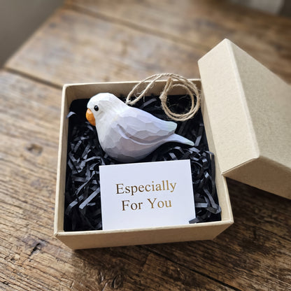 A white Lovebirds ornament with a small orange beak nestled in black shredded paper inside a kraft gift box, with a card reading 'Especially For You' and a twine loop above the bird.