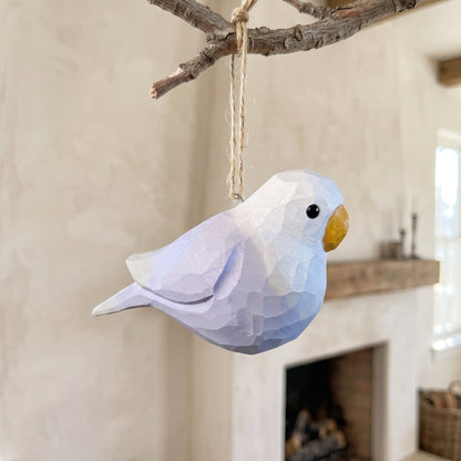 A low-poly Lovebirds ornament in light blue and white hangs from a natural branch with twine in a softly blurred indoor setting.