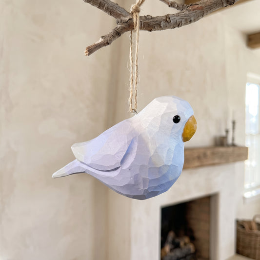 A low-poly Lovebirds ornament in light blue and white hangs from a natural branch with twine in a softly blurred indoor setting.