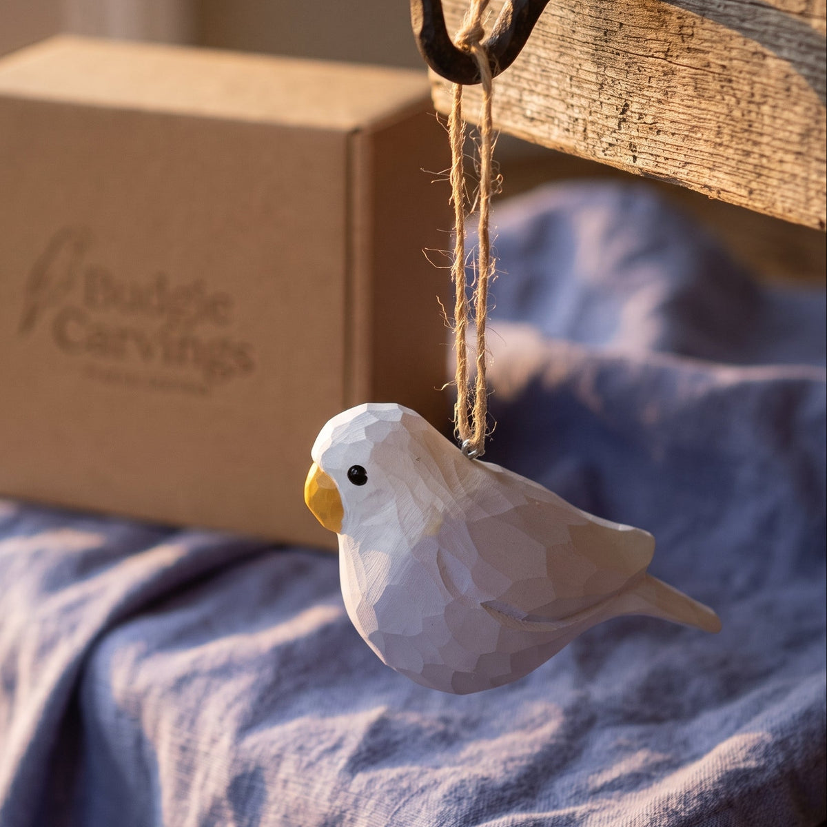 A white faceted wooden Lovebirds hanging ornament with a yellow beak, suspended by twine from a rustic wooden hook. A brown cardboard box with the Budgie Carvings logo sits in the background on a blue cloth.