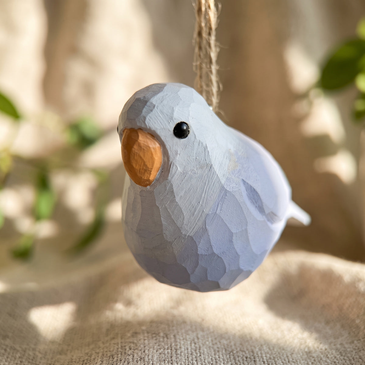 A small wooden Lovebirds hanging ornament with a faceted geometric design, painted in pale blue-gray with a natural light brown wood beak, hanging from a twine string against a soft-focus beige background.