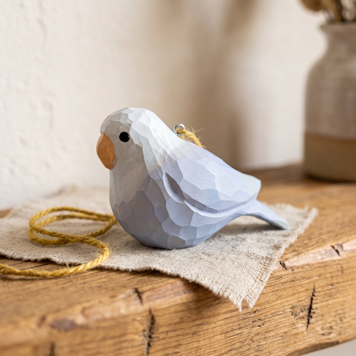 A small Lovebirds-shaped hanging ornament with a faceted geometric design, painted in soft light blue and white, with a natural wood beak and a yellow hanging string, resting on a piece of burlap over a rustic wooden surface.