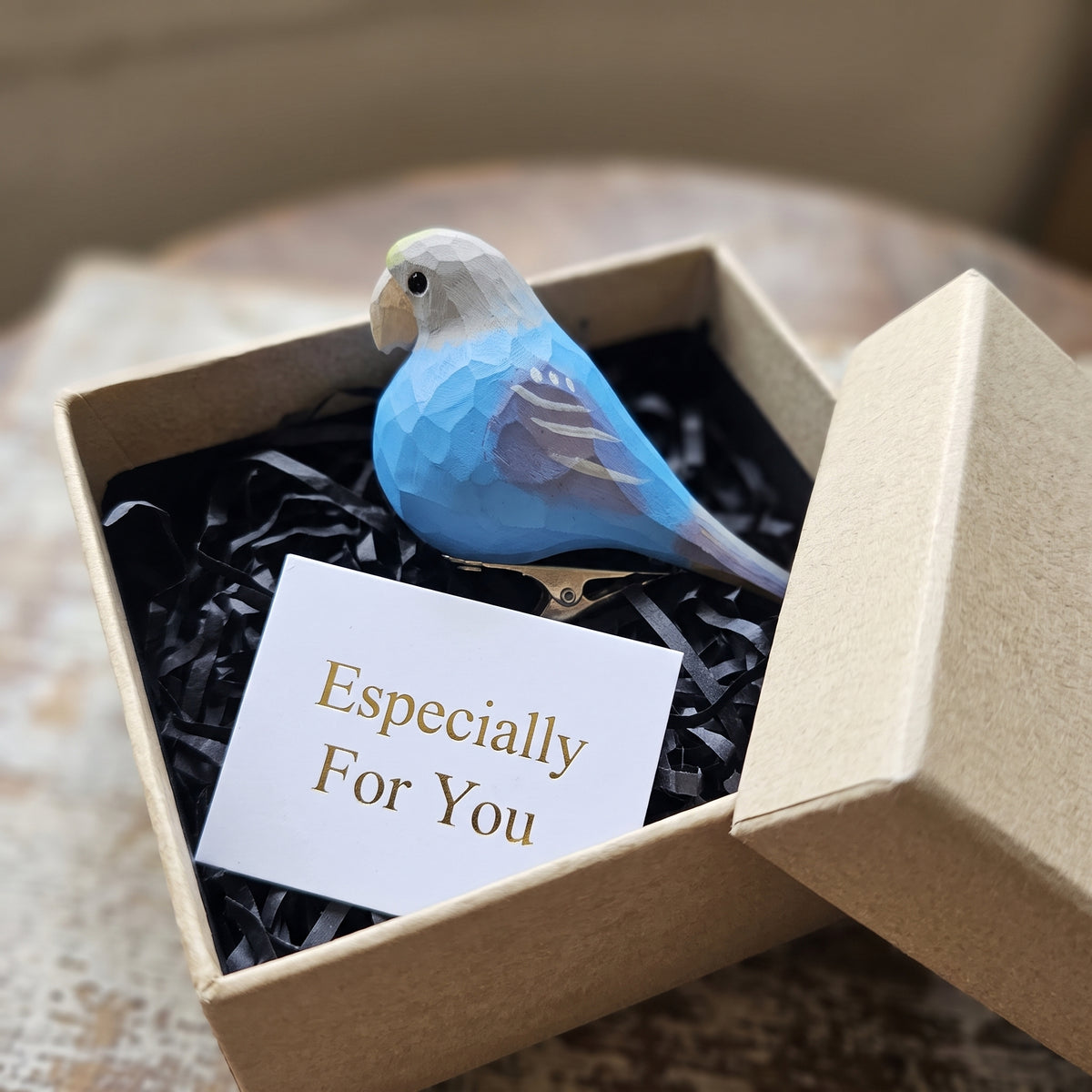 A hand-carved blue wooden lovebird clip-on ornament resting in an open kraft gift box filled with black shredded paper, alongside a small card reading "Especially For You".