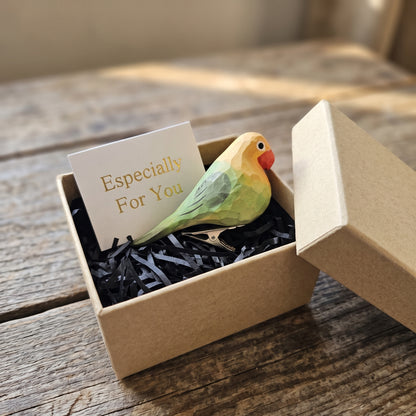 A hand-carved pastel green wooden lovebird clip-on ornament resting in an open kraft gift box filled with black shredded paper, alongside a small card reading "Especially For You" and the box lid propped beside it on a rustic wooden surface.