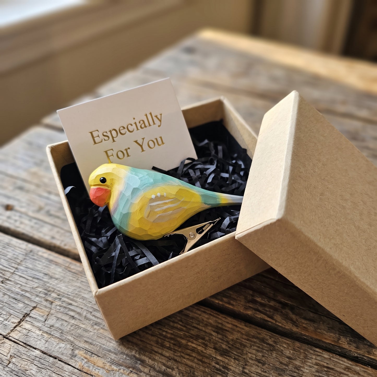 A hand-carved wooden lovebird clip-on ornament resting in an open kraft gift box filled with black shredded paper, featuring pastel yellow, green, pink, and turquoise-blue painted plumage, a light red beak, a silver metal clip attached underneath, and a small card reading "Especially For You" placed behind it.