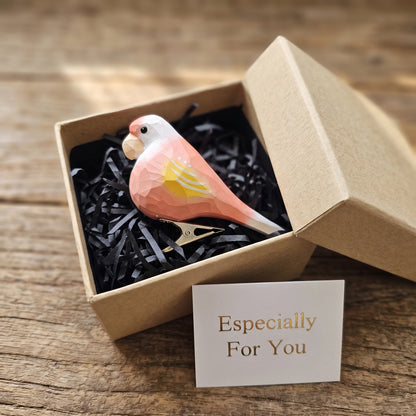 A hand-carved pink wooden lovebird clip-on ornament resting in an open kraft gift box filled with black shredded paper, featuring a pastel pink body, yellow and white wing accents, a light orange beak, and a small card reading "Especially For You" placed in the foreground.