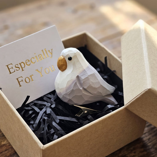 A hand-carved wooden white lovebird figurine with a yellowish beak resting inside an open kraft paper gift box filled with black shredded paper, alongside a white card reading "Especially For You" in the foreground on a rustic wooden surface.