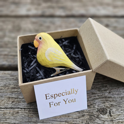 A hand-carved yellow wooden lovebird clip-on ornament resting in an open kraft gift box with black shredded paper, featuring a light orange beak, a silver metal clip attached underneath, and a white card reading "Especially For You" placed in the foreground.