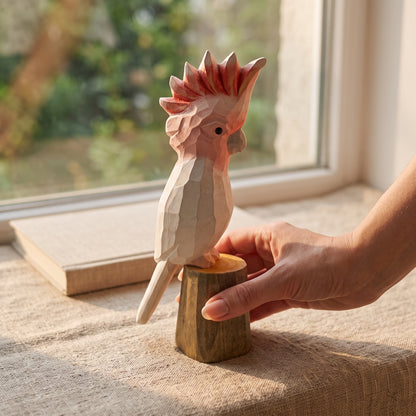 Hand holding a hand-carved wooden Pink Cockatoo bird figurine by its stump base on a sunlit table near a window.