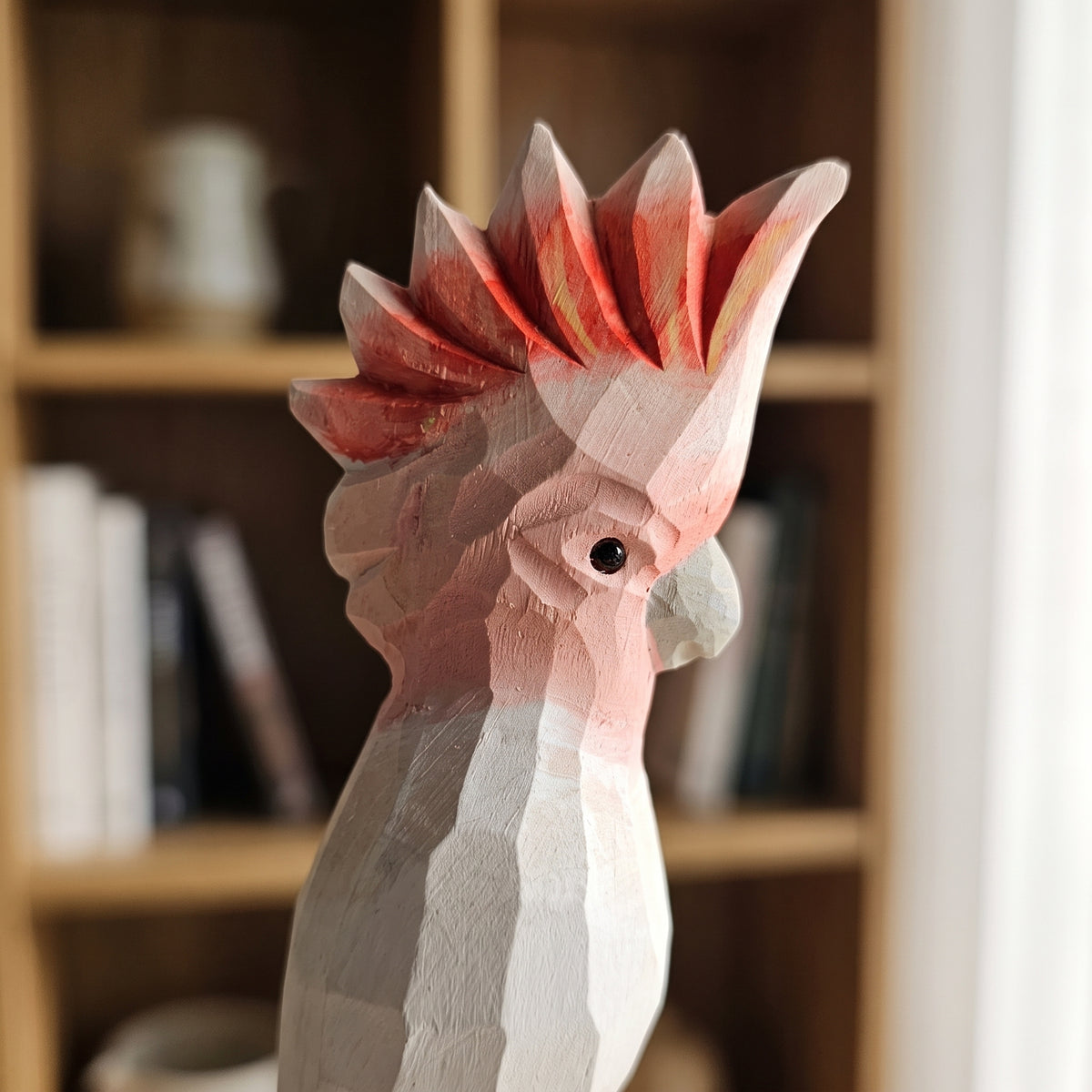 Close-up of a hand-carved wooden Pink Cockatoo bird head showing faceted details, black eye, and fan-shaped crest, with a softly blurred bookshelf background.