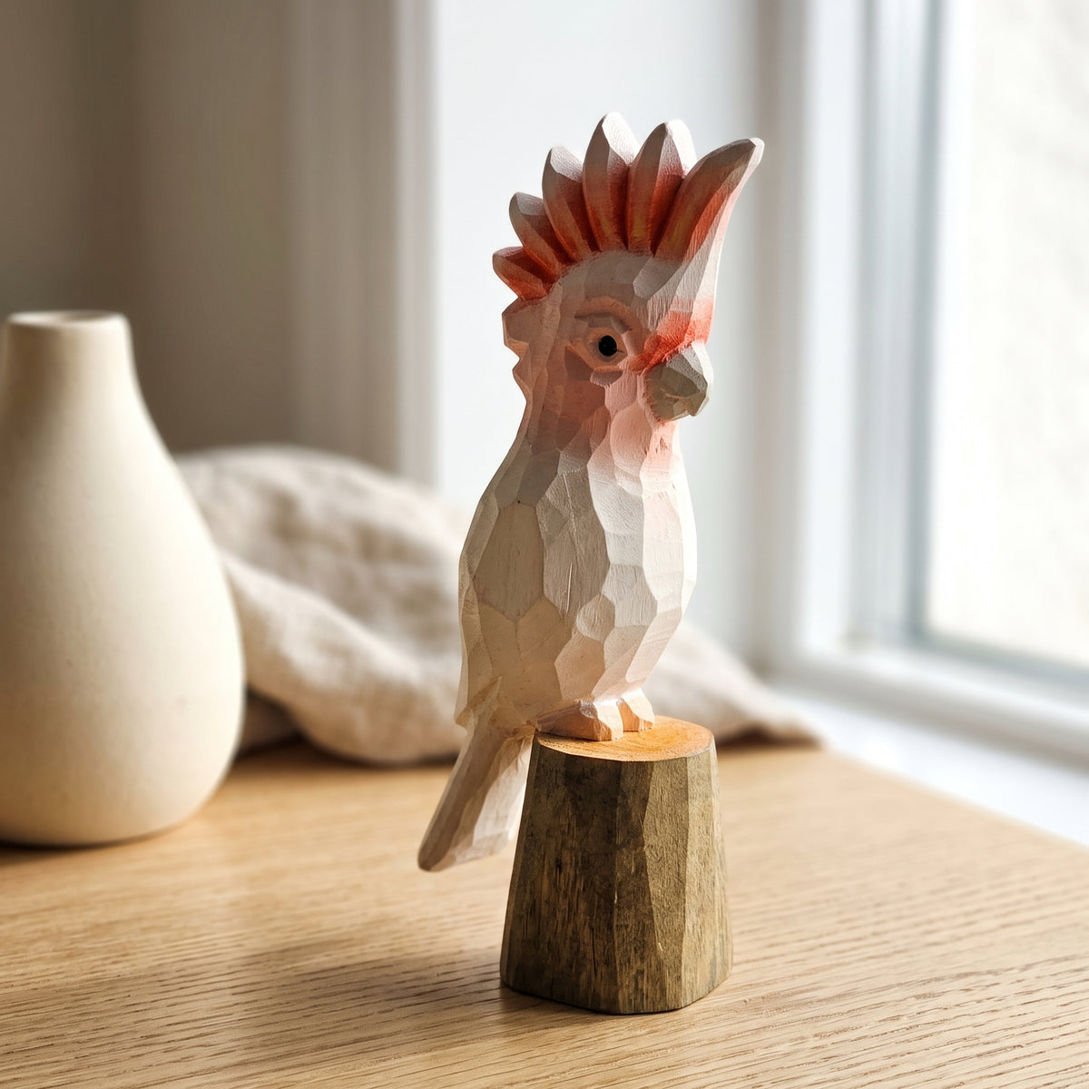 Hand-carved wooden Pink Cockatoo bird figurine with a pink crest, perched on a wooden stump base on a light wooden table near a window.