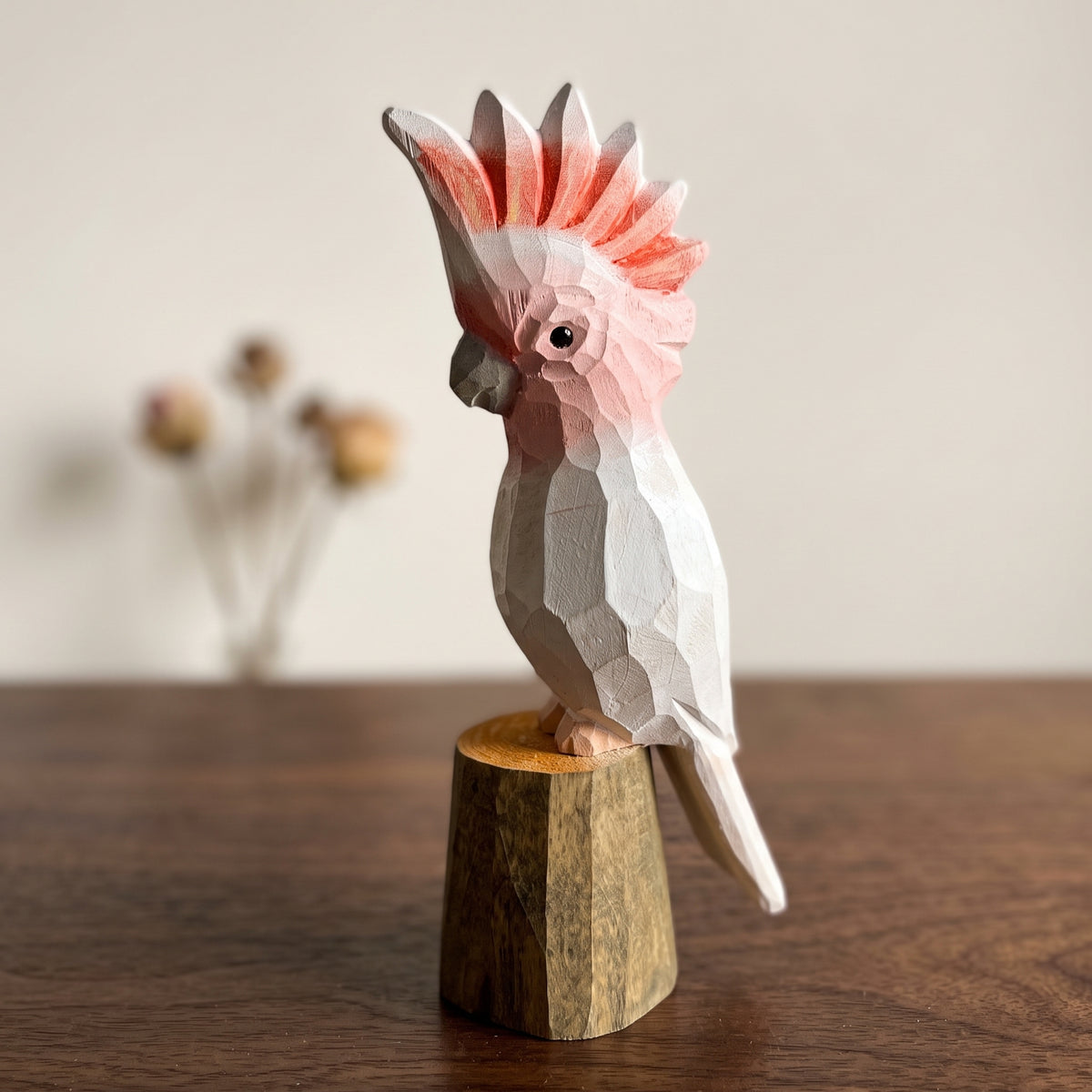 Hand-carved wooden Pink Cockatoo bird figurine with a pink and white crest, perched on a small wooden stump base on a dark tabletop.