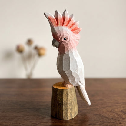 Hand-carved wooden Pink Cockatoo bird figurine with a pink and white crest, perched on a small wooden stump base on a dark tabletop.