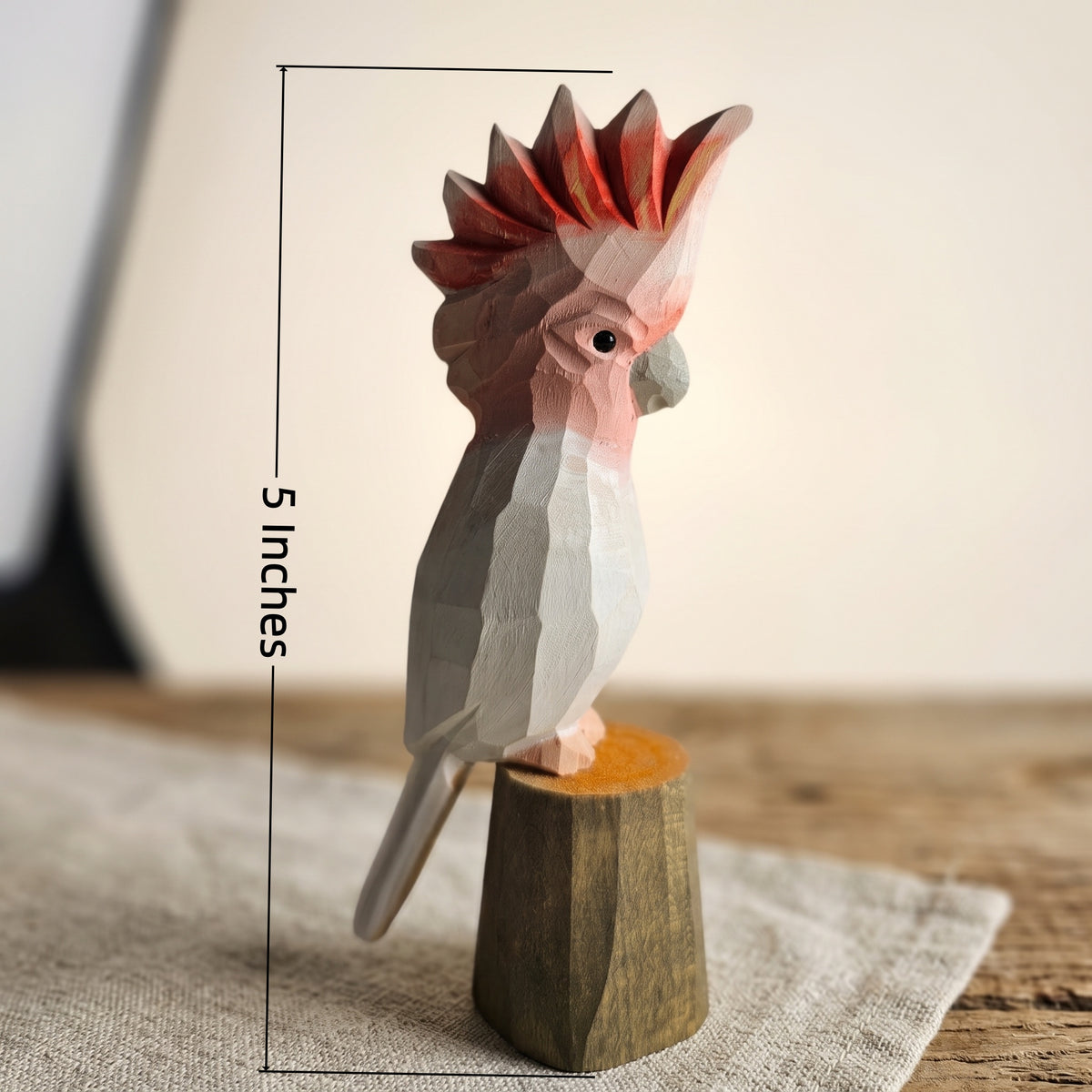 Size graphic showing a 5-inch hand-carved wooden Pink Cockatoo bird figurine perched on a wooden stump base on a linen tablecloth.
