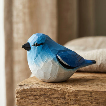 A hand-carved wooden Mountain bluebird figurine painted in shades of blue and white, with a black beak, resting on a rustic wooden block with a soft-focus neutral background.