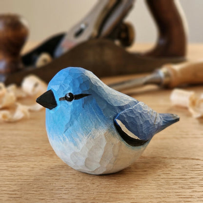 A small hand-carved wooden Mountain bluebird figurine painted blue and white, placed on a wooden surface with woodworking tools blurred in the background.
