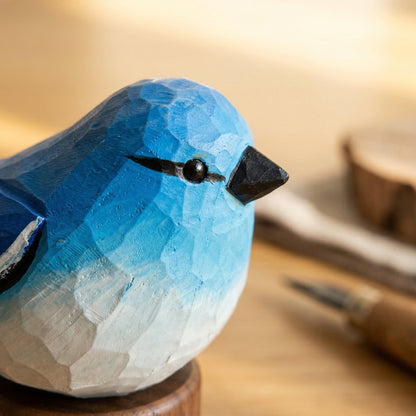 A close-up of a hand-carved wooden Mountain bluebird figurine with a blue and white gradient finish, resting on a wooden surface with a blurred background.