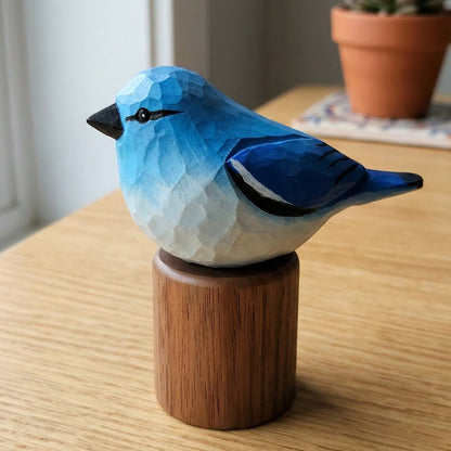 A small Mountain bluebird figurine with a faceted geometric texture, painted in shades of blue and white, perched on a cylindrical wooden base. The figurine is set on a light wood table in a home environment with a potted succulent in the background.