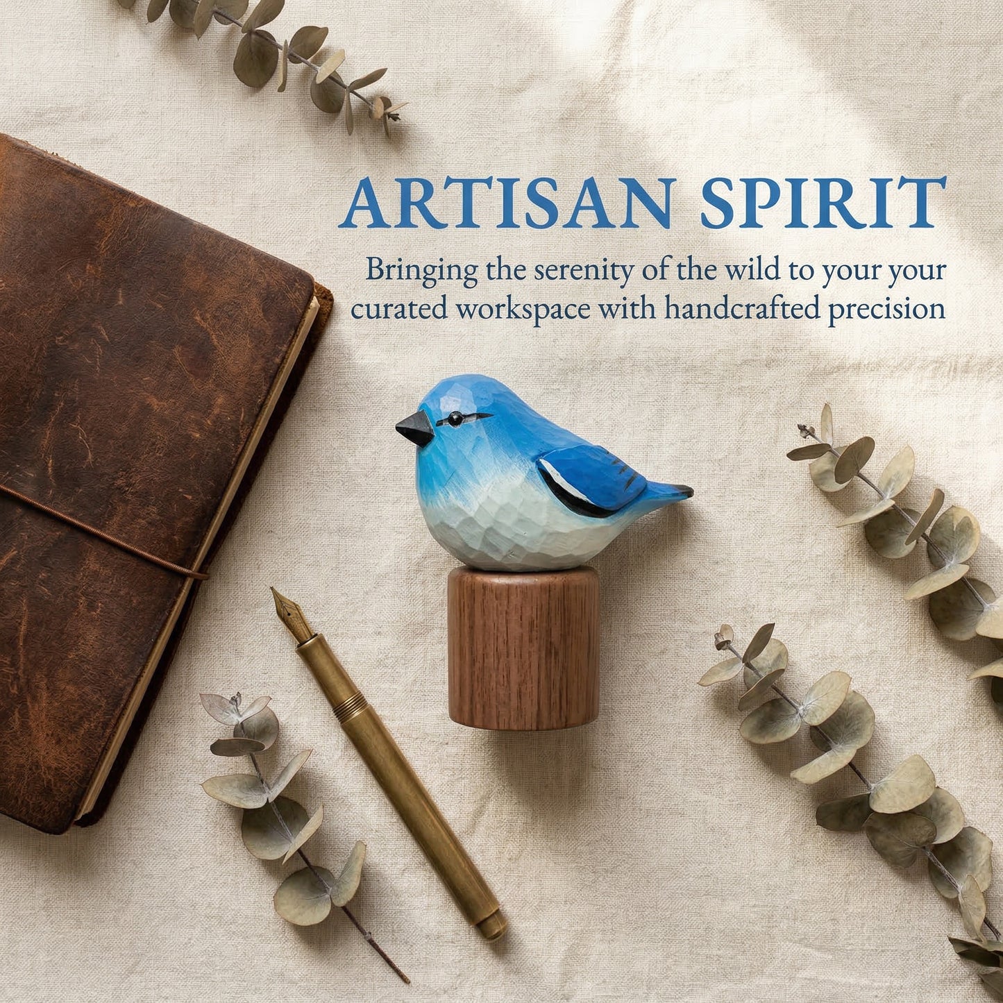 A small Mountain bluebird  figurine painted with geometric facets, mounted on a round wooden base, styled on a beige fabric surface with a brown notebook, gold pen, and eucalyptus sprigs. Blue text reads 'ARTISAN SPIRIT' with a subtitle about bringing the serenity of the wild to a curated workspace.