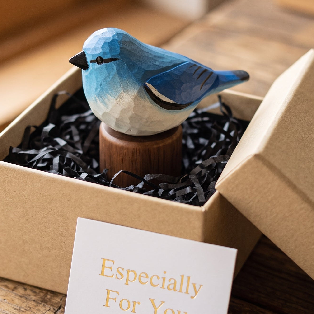 A small Mountain bluebird figurine displayed in an open kraft gift box with black shredded paper filler and a white card reading 'Especially For You' on a rustic wooden table.