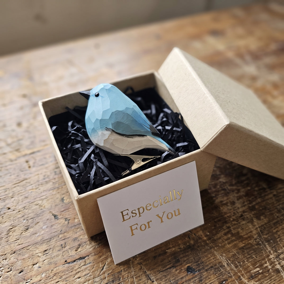 A hand-carved light-blue wooden mountain bluebird clip-on ornament resting in an open kraft gift box filled with black shredded paper, alongside a small card that reads "Especially For You".