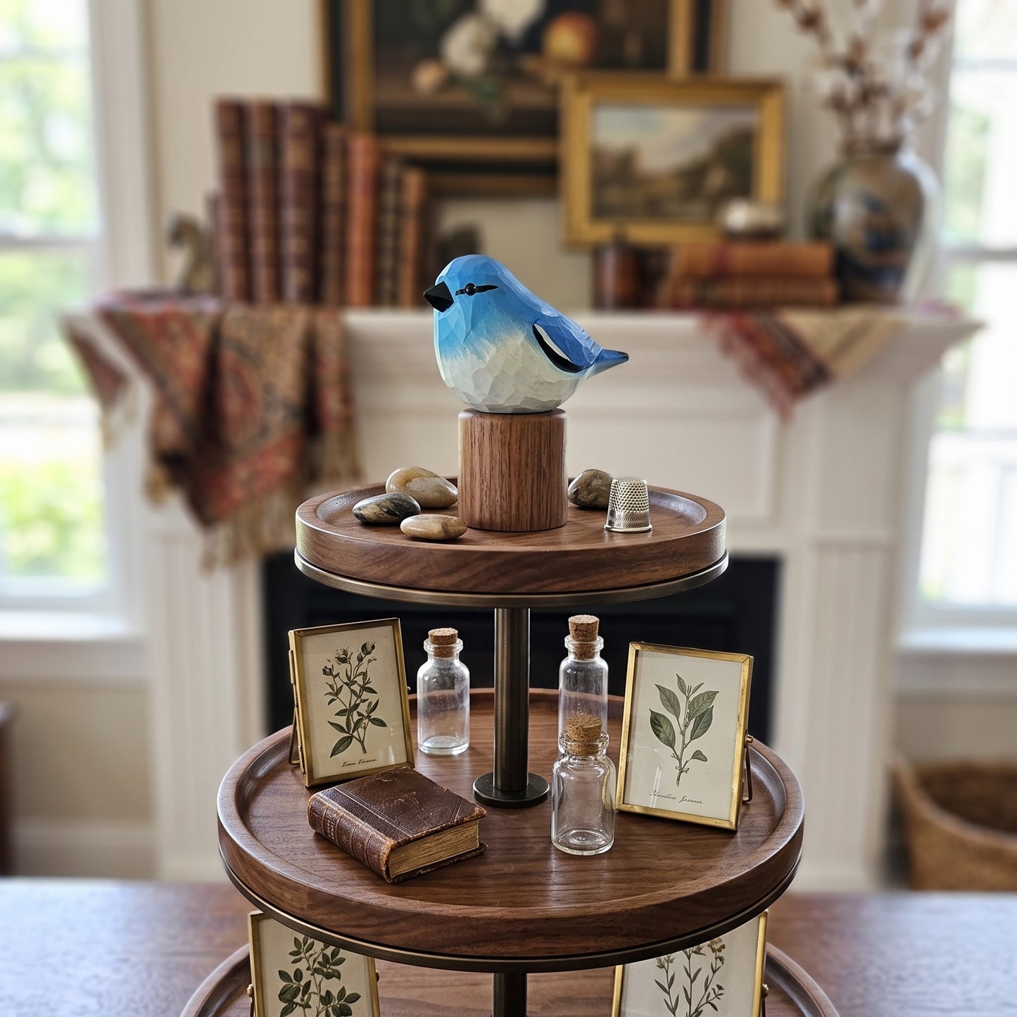 A three-tier wooden display stand styled with miniature books, small glass bottles, framed botanical prints, and a Mountain bluebird figurine on a round wooden top, set in a cozy, softly blurred room with a fireplace and windows in the background.