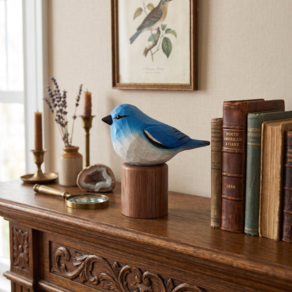 A blue and white Mountain bluebird figurine perched on a cylindrical wooden base, displayed on a carved wooden console table in a styled room setting with books, candles, and framed wall art.