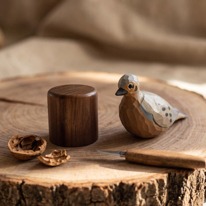 A handcrafted wooden Mourning Dove figurine with a matching dark wooden cap sits on a rustic wood slice next to a small carving knife and walnut shells.